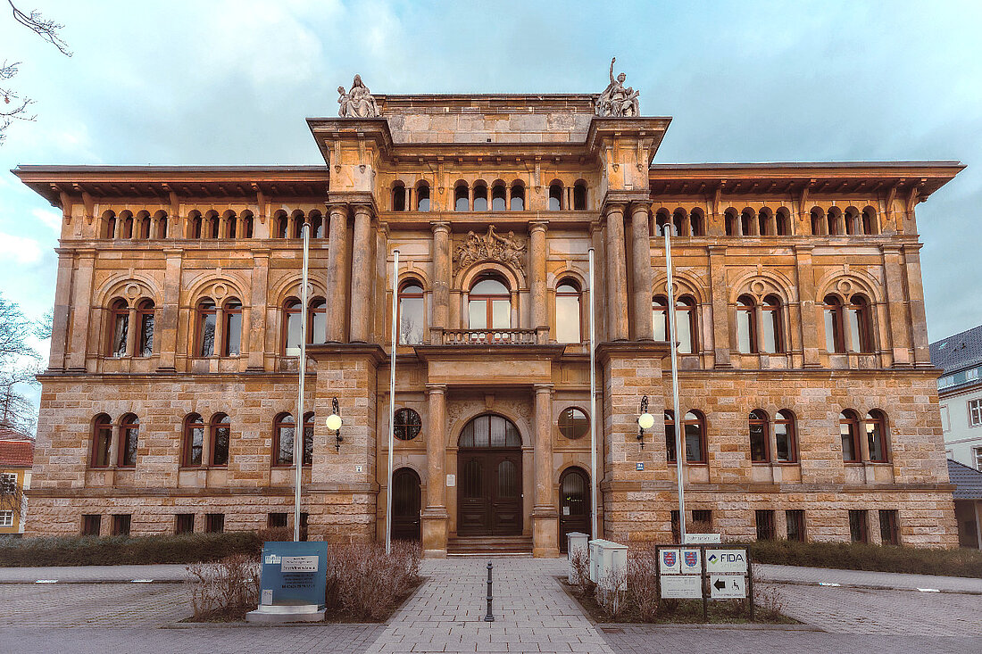 Foto des Gebäudes. Man sieht das historische Gebäude aus Sandstein mit Flachdach sowie einen Teil des Parkplatzes.